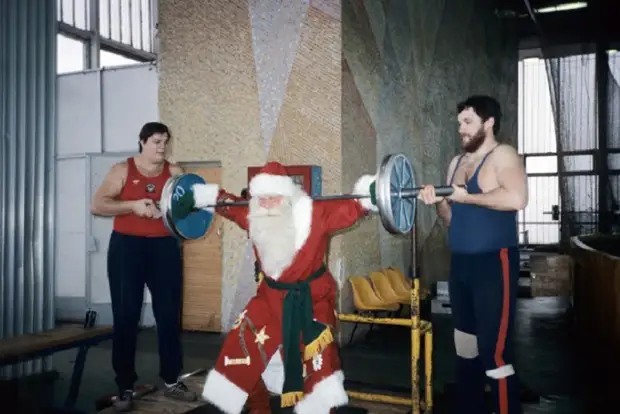 Дед Мороз пытается поднять спортивный снаряд, под чутким руководством спортсменов.