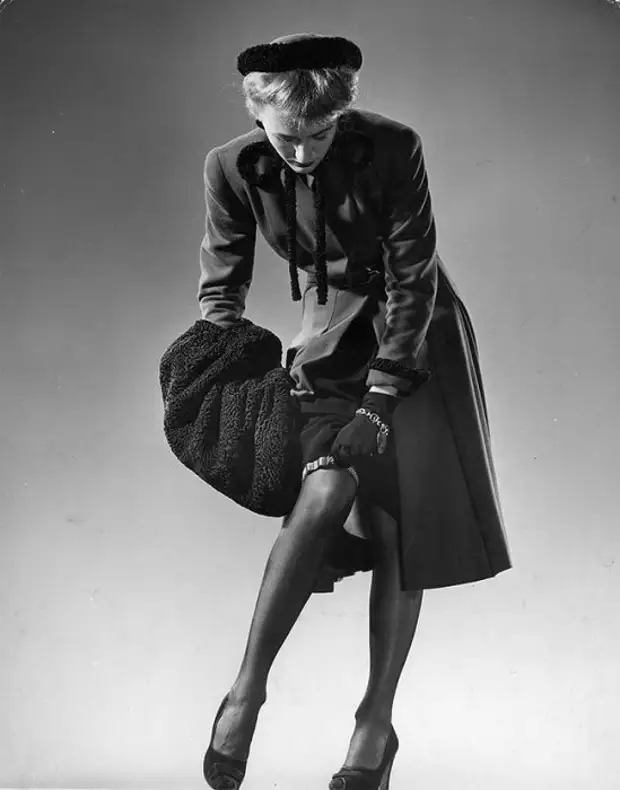 Модель в черных чулках, приталенном черном платье, меховой шляпке и с меховой муфтой поправляет подвязки, 1946 год.
